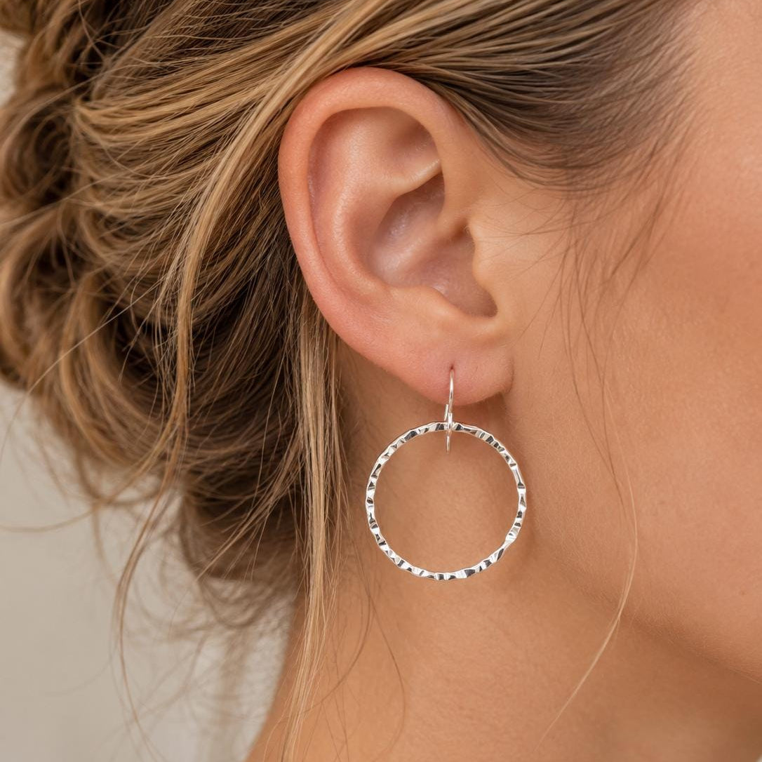 A close-up of a woman's ear wearing a textured silver hoop earring with a small silver fishhook back.
